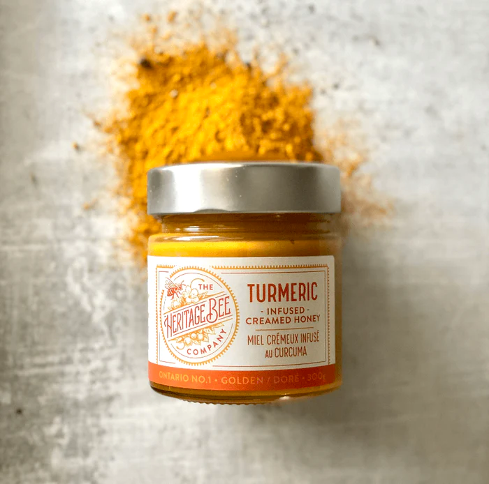 A jar of Elderberry honey from The Heritage Bee Company, surrounded by decorative flowers and a honey dipper on a white plate.