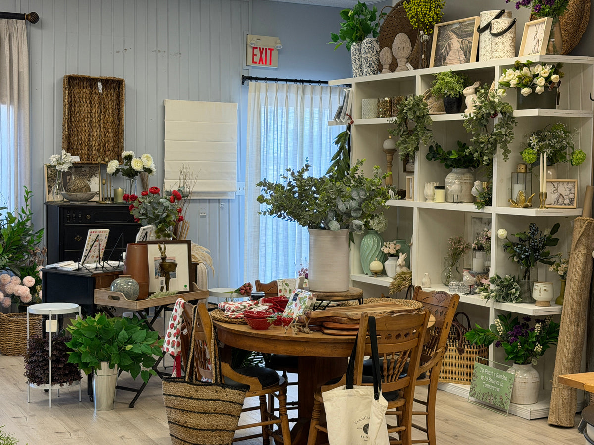A styled interior room with a round wooden dining table and chairs in the foreground, surrounded by shelves filled with potted plants, vases, baskets, and home decor. A whiteboard, curtains, and an exit sign are visible in the background.