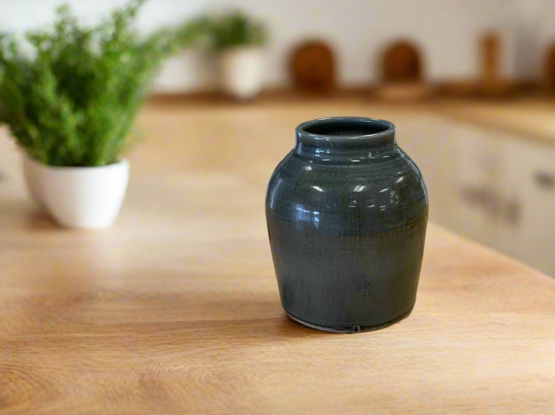 A grey ceramic pot placed on a wooden surface, likely a tabletop, with a plant in the background.