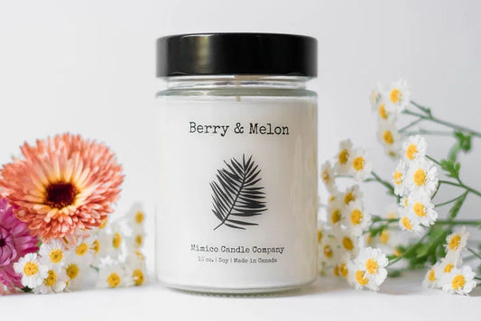 A soy wax candle in a clear glass jar with a black lid, labeled 'Decor Chic Living' and the scent 'Satsuma'. The candle is displayed on a wooden surface with greenery in the background.