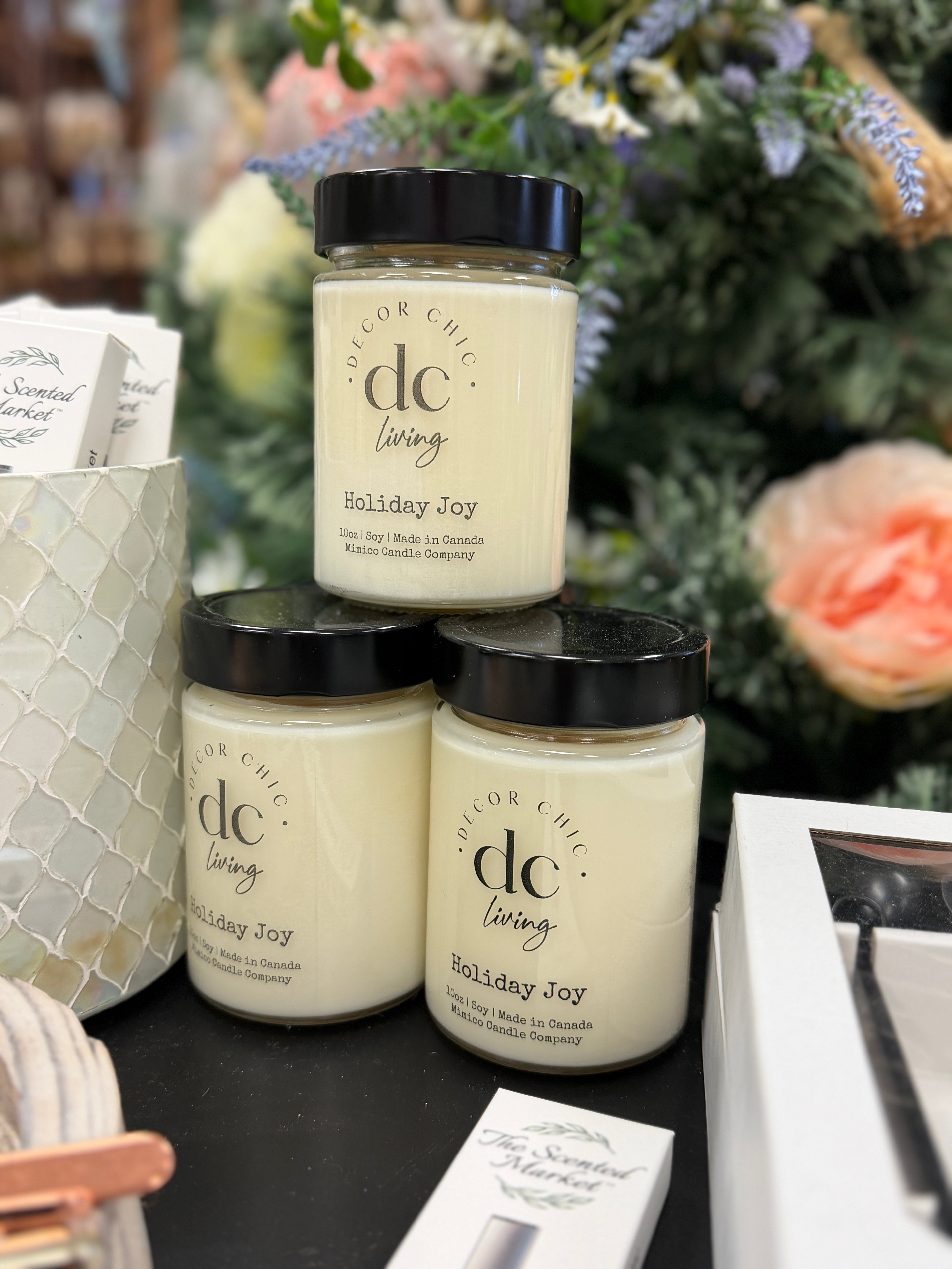 Three jars of 'dc living' Holiday Joy candles stacked on a table with decorative background.