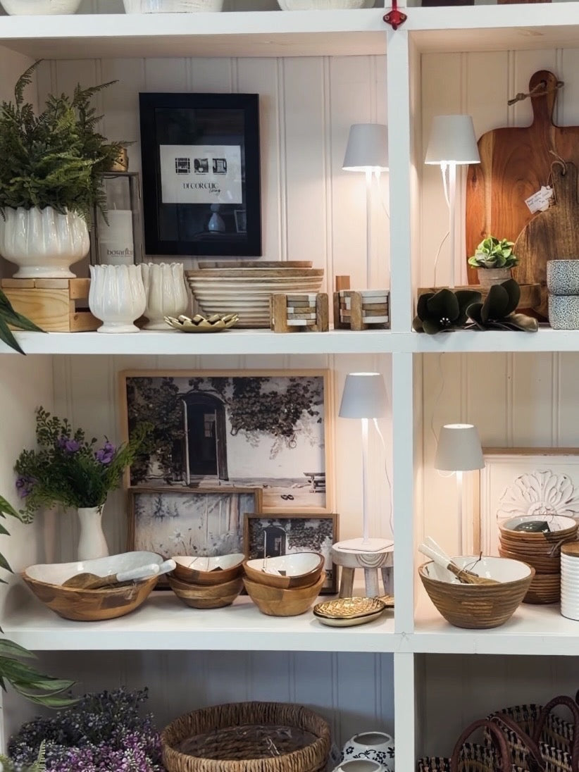 A styled interior shelf display with white shelving holding ceramic vases, bowls, framed wall art, small lamps, baskets, and decorative plants in a warm home decor setting.