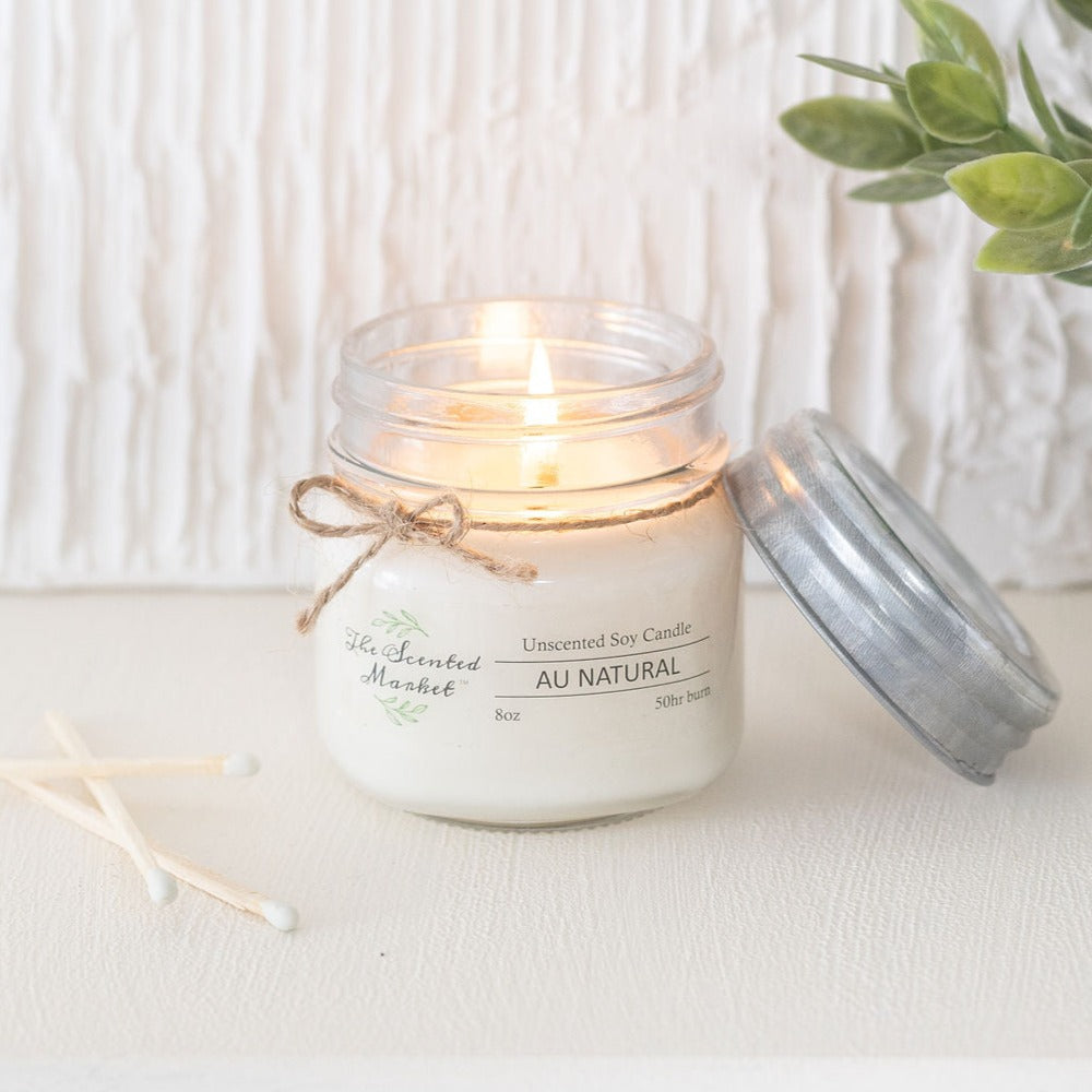 Unlit candle in a glass jar with a label on a white surface, accompanied by a plant.