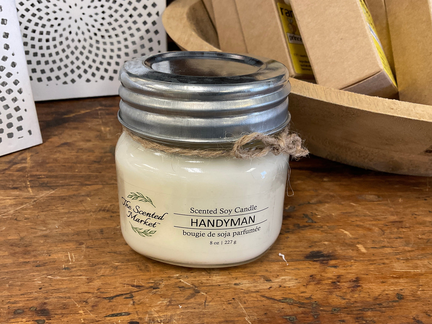 Scented soy candle labeled 'Handyman' in a mason jar on a wooden surface with cardboard boxes in the background.