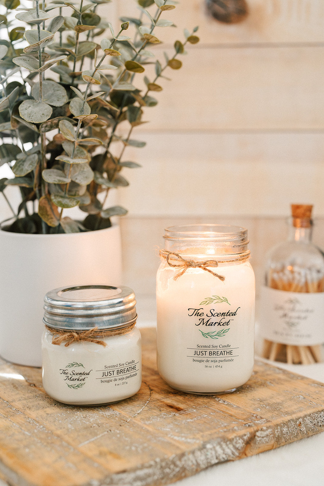 Two candles with decorative bows on a wooden surface, surrounded by plants and bottles.