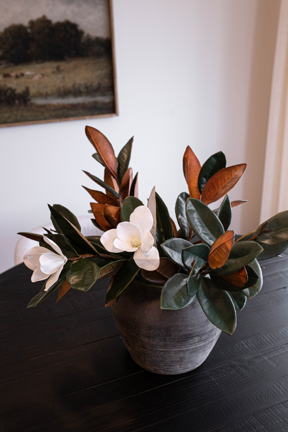 Small faux magnolia stems arranged in a dark vase on a dining table in a neutral interior