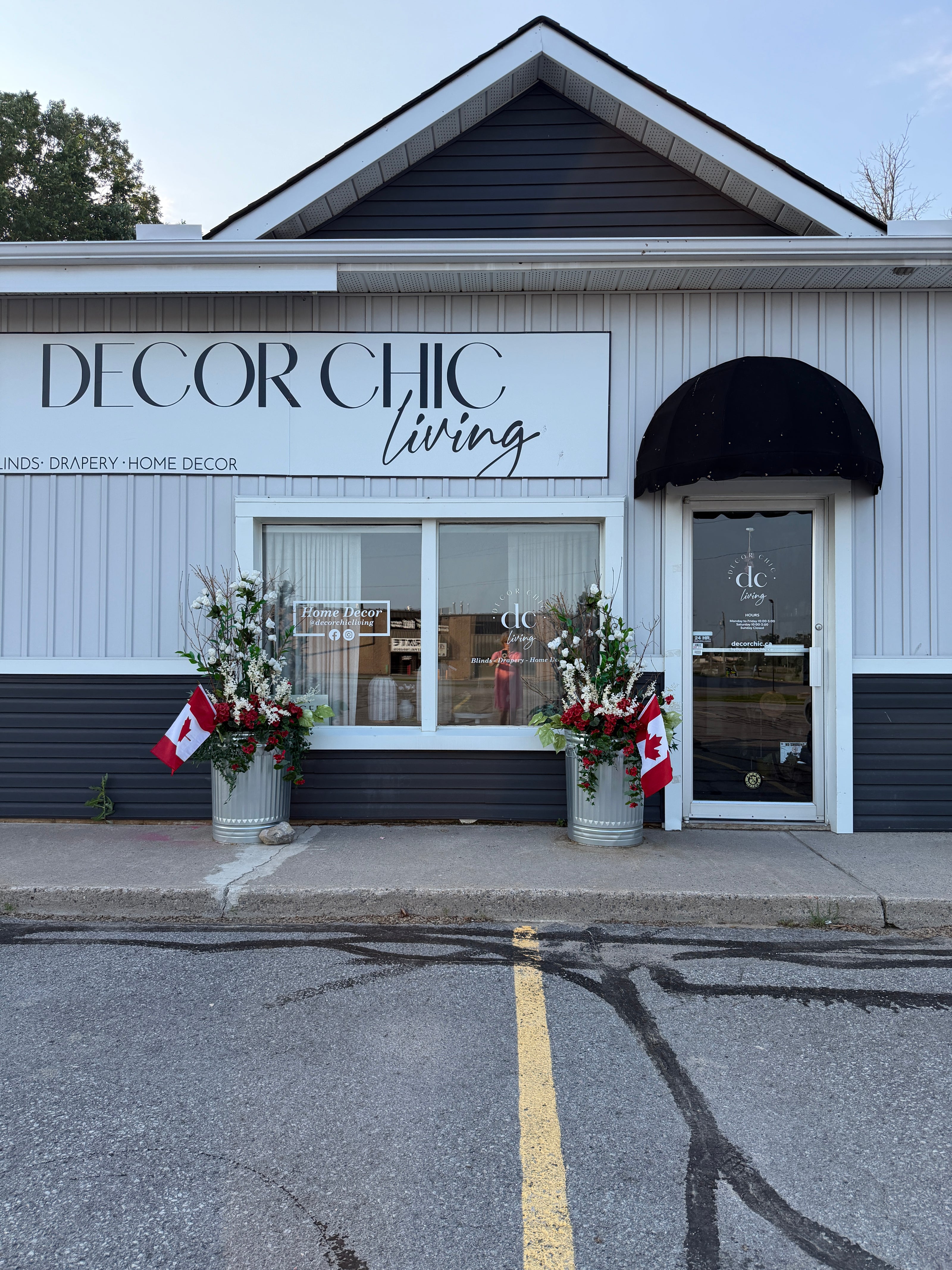 Decor Chic Living storefront on a quiet street, featuring white siding, dark gray trim, a black arched awning, large windows, and two flower arrangements with Canadian flags on the sidewalk.