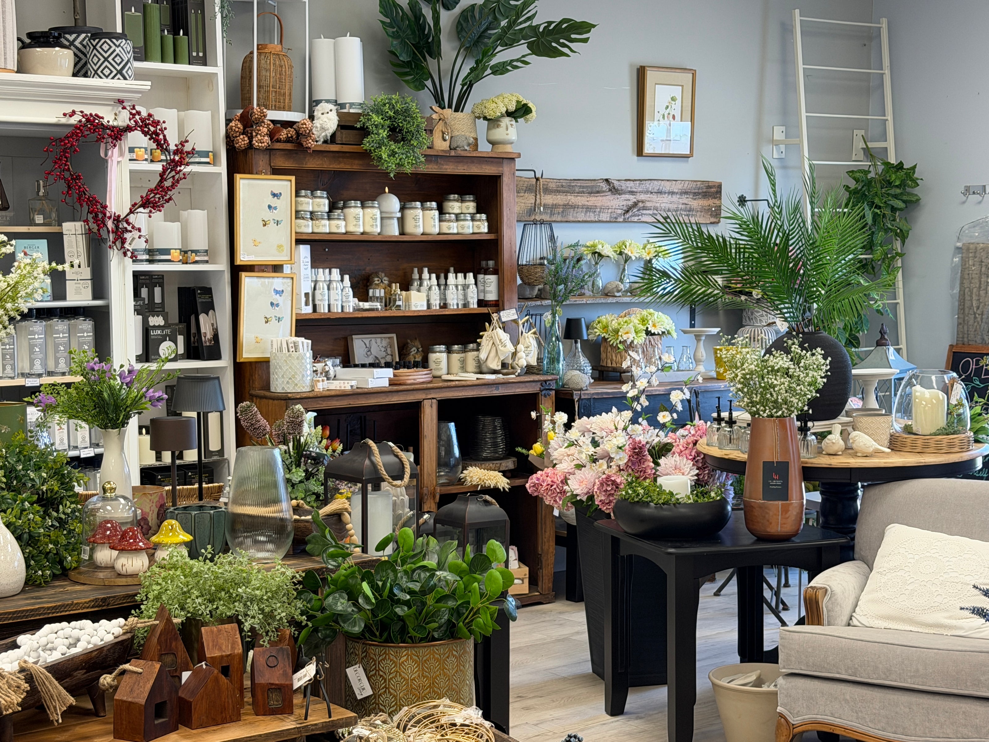 A cozy, rustic home decor and gift shop interior filled with plants, vases, candles, wooden shelves, and floral arrangements in a bright, styled room.